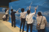 屋内の会場にて、数名の男女が横に並び、壁一面に張られた巨大な青いボードに向かって、筆のような道具を使いながら白い雲の模様を描き込んでいる写真