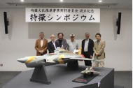 「特撮シンポジウム」と書かれた横断幕の下で、精巧な戦闘機の大型モデルが展示された後方で、スーツやカジュアルな服装の男性6名が並んだ記念写真