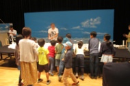 屋内の会場にて、青空が描かれた巨大な背景パネルの前で、多くの子供たちや大人が集まり、スタッフが中央で解説を行っている様子を捉えた写真