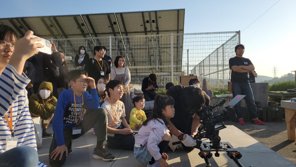 屋外で、本格的なビデオカメラで撮影しているスタッフと、子どもたちが興味深そうに撮影の様子を近くで見守っている写真
