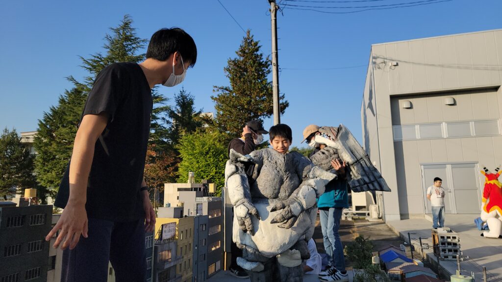 青空の下、ビルが立ち並ぶ精巧なミニチュアセットの中で、岩のような質感の怪獣の着ぐるみを身にまとった少年をスタッフたちが囲み、撮影の準備や演技の調整を丁寧に行っている特撮現場の様子を捉えた写真