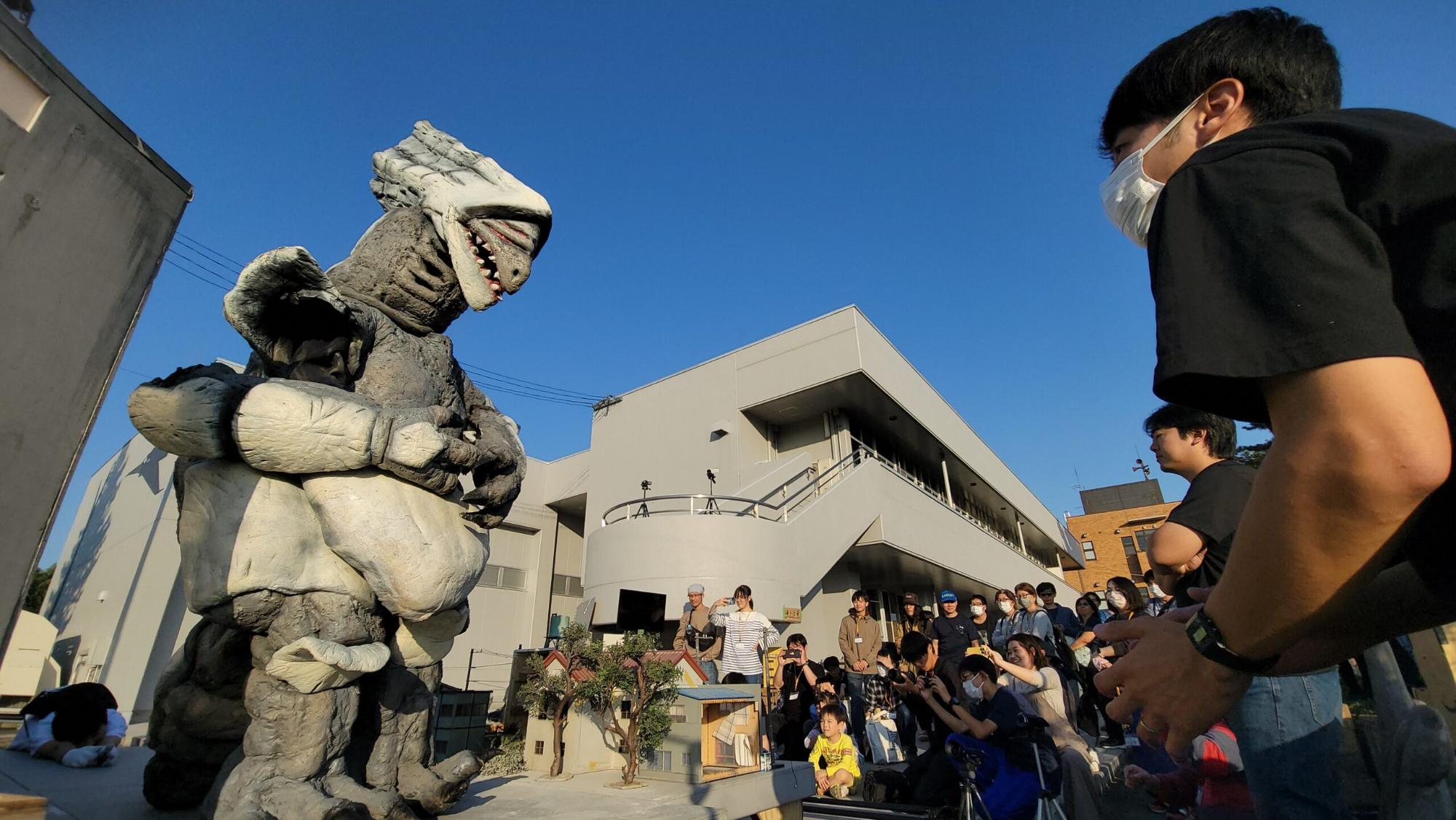 屋外の撮影現場にて、灰色の怪獣の着ぐるみがミニチュアの民家の前に立ち、その様子を多くのスタッフや見学者がスマートフォンなどを手に持ちながら囲んで見守っている風景を収めたローアングルの写真