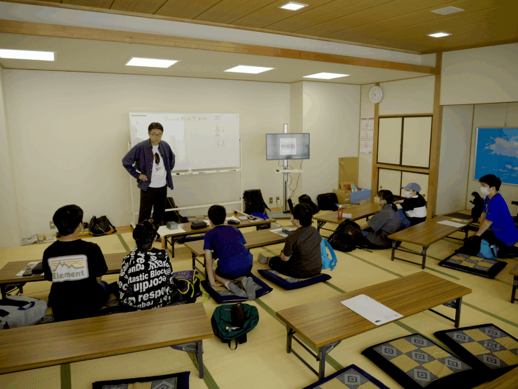 座卓と座布団が並ぶ和室で講師がホワイトボード前に立ち教材や画面を用いて受講者に説明している様子の写真