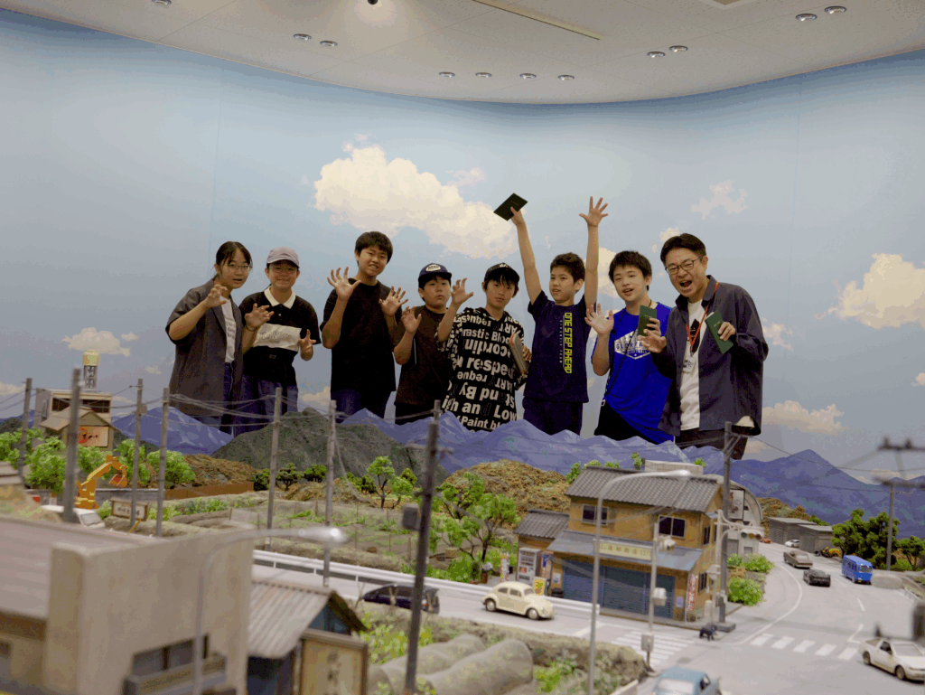 青空と白い雲が描かれたホリゾントを背景に、山並みや民家、道路が再現された精巧なジオラマセットの向こう側で、笑顔の学生たちと講師が怪獣のポーズをとったり手を挙げたりして並んでいる、特撮スタジオでの記念写真