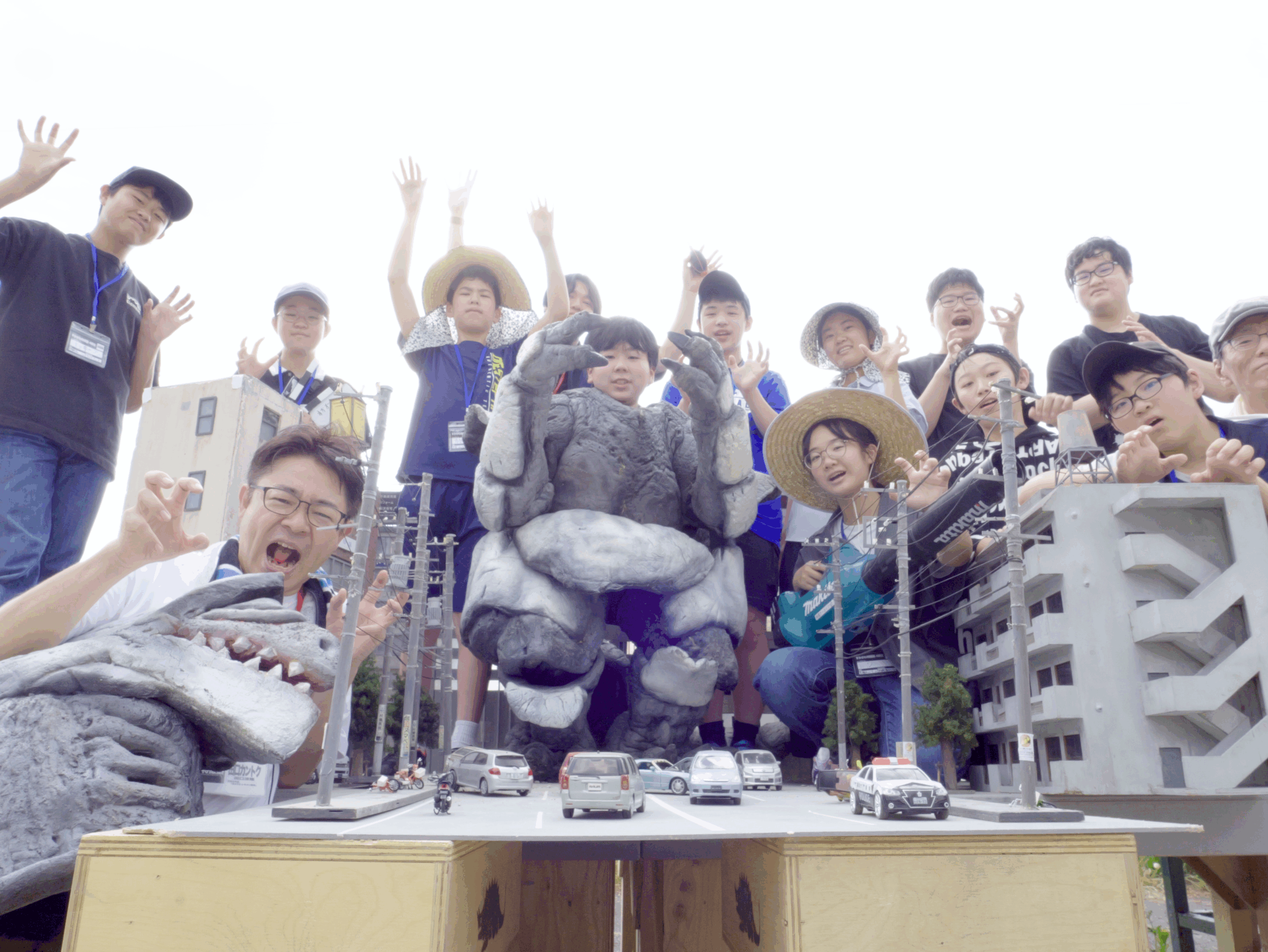 屋外の特撮セットで、手作りのビル模型やミニチュアカーを囲み、中央に岩のような怪獣、左端に恐竜型の怪獣を配置して、大勢の学生たちが怪獣になりきったポーズで笑顔を見せている、特撮ワークショップの集大成となる集合写真