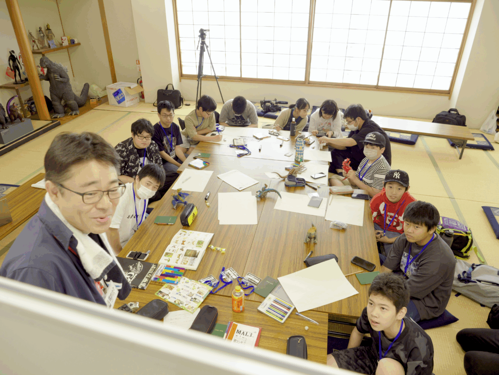 明るい光が差し込む和室で、中央の大きな長机を囲んだ学生たちが、手元のワークブックや資料を広げながら真剣に講師の話を聞いたり作業を進めたりしている、特撮ワークショップの座学風景を捉えた写真