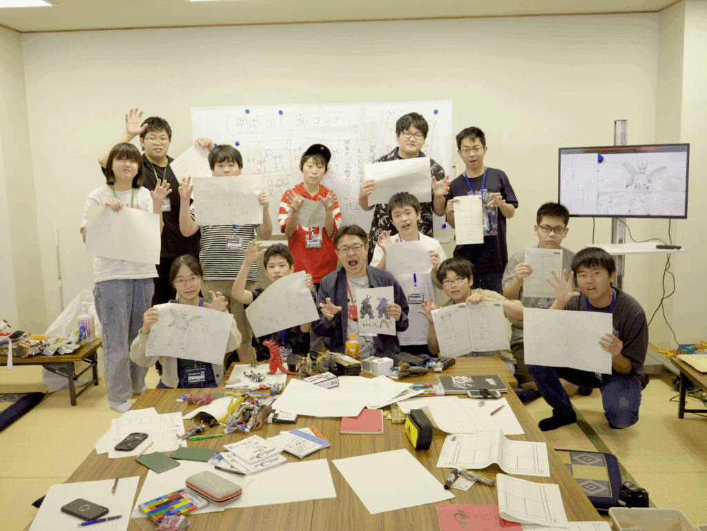 多数の参加者たちが完成させた怪獣などのイラストを高く掲げ、資料や画材が散らばる座卓の前で達成感に満ちた記念撮影を行う写真