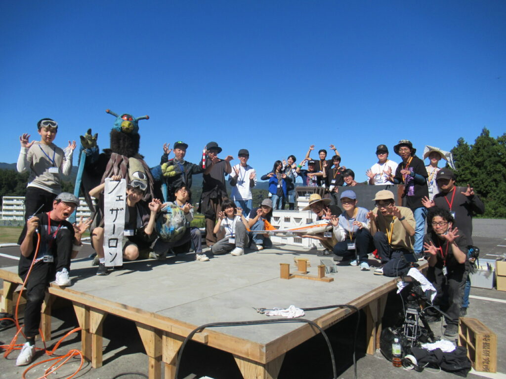 青空の下、屋外の木製台上に集まった大人数が怪獣の着ぐるみとともに手を広げてポーズを取り楽しげに記念撮影する写真