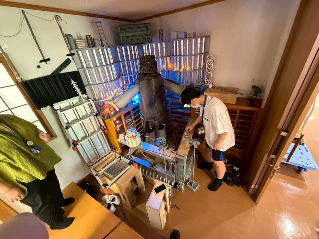 巨大なロボット風オブジェとビル群の精巧なセットの前で参加者が装置を調整する様子の写真