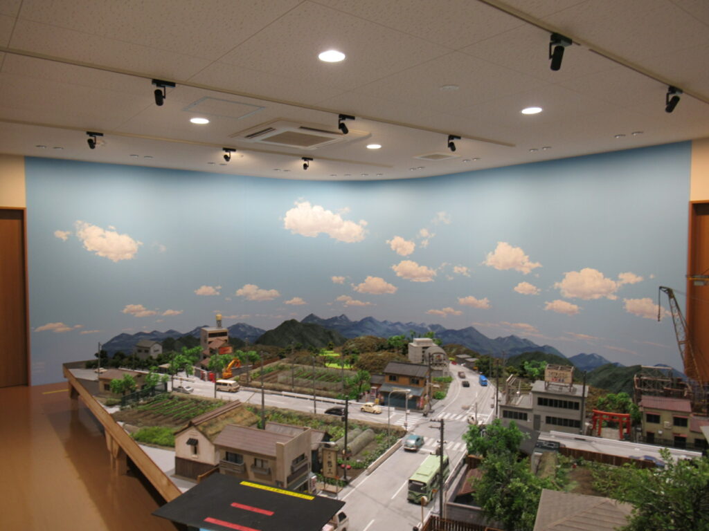 青い空と白い雲が描かれた背景の前に、山々や家屋、道路、走るバスなどが精巧に再現された街並みの巨大なミニチュアセットの全体を写した写真