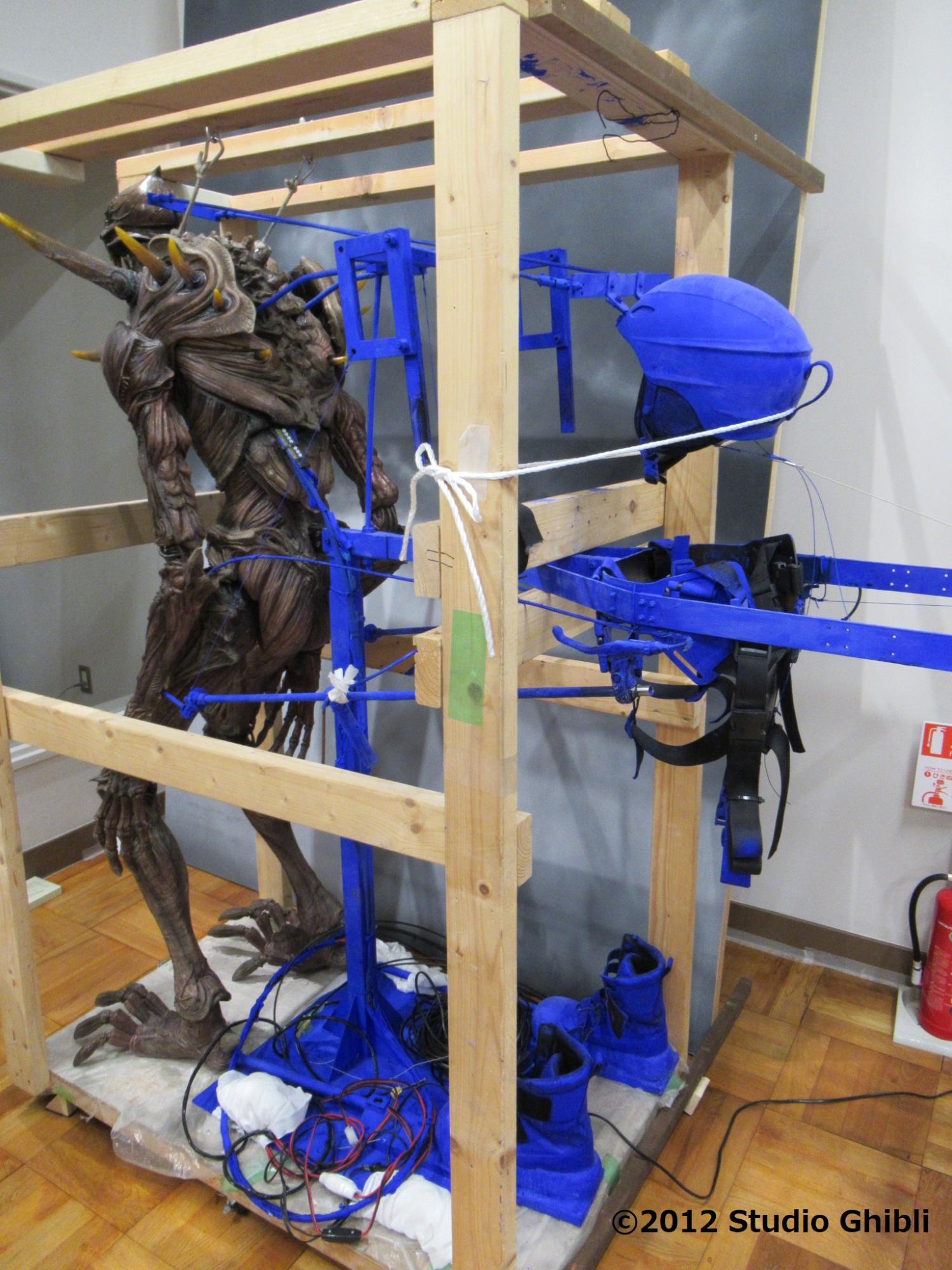 木製のフレーム内に固定された、筋繊維が剥き出しのような茶色い身体を持つ巨神兵の全身像の背面に、青い支持具や操作用のフレーム、ヘルメット、ブーツなどの操演用機材が取り付けられた状態を背後から捉えた写真
