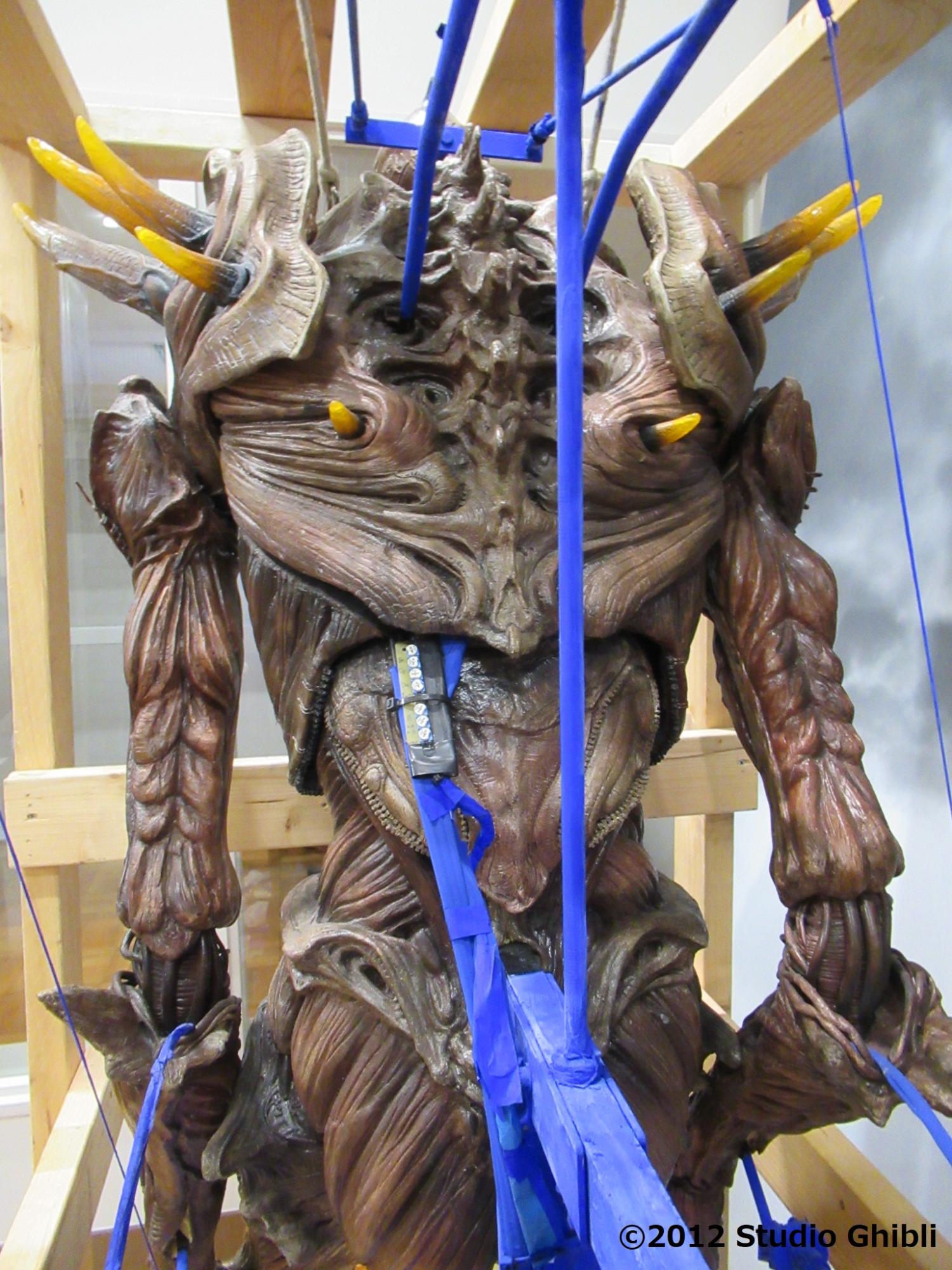 特撮短編映画に登場する巨神兵の背後からのクローズアップで、有機的な質感を持つ茶色い背甲に、青い操作用のロッドや支持具が直接取り付けられ、周囲を木枠に囲まれた写真