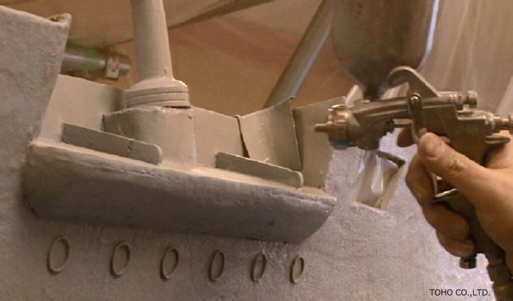 人の手によってスプレーガンが握られ、グレーに塗装された戦艦のミニチュア模型の細部へと精密に塗料が吹き付けられている写真