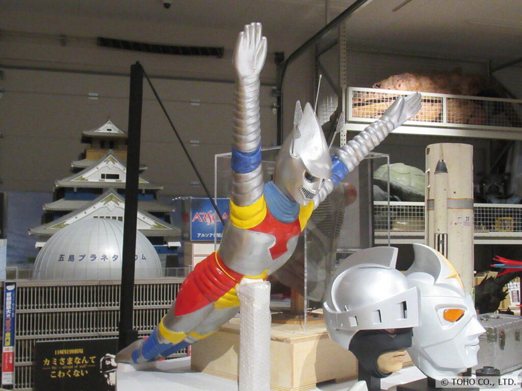 特撮ヒーローが両腕を突き出し飛行するポーズのフィギュアを中心に、手前にはヒーローのマスク、背景にはお城の模型やプラネタリウムのドームが並ぶ、特撮作品の資料や小道具を一同に集めた展示写真
