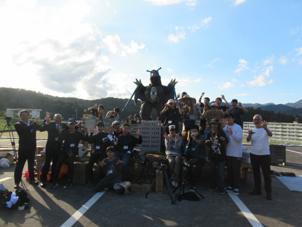 晴天の屋外にて、精巧なミニチュアの街並みを背景に怪獣の着ぐるみを囲んで全員でポーズを決めるスタッフたちの姿を捉えた集合写真