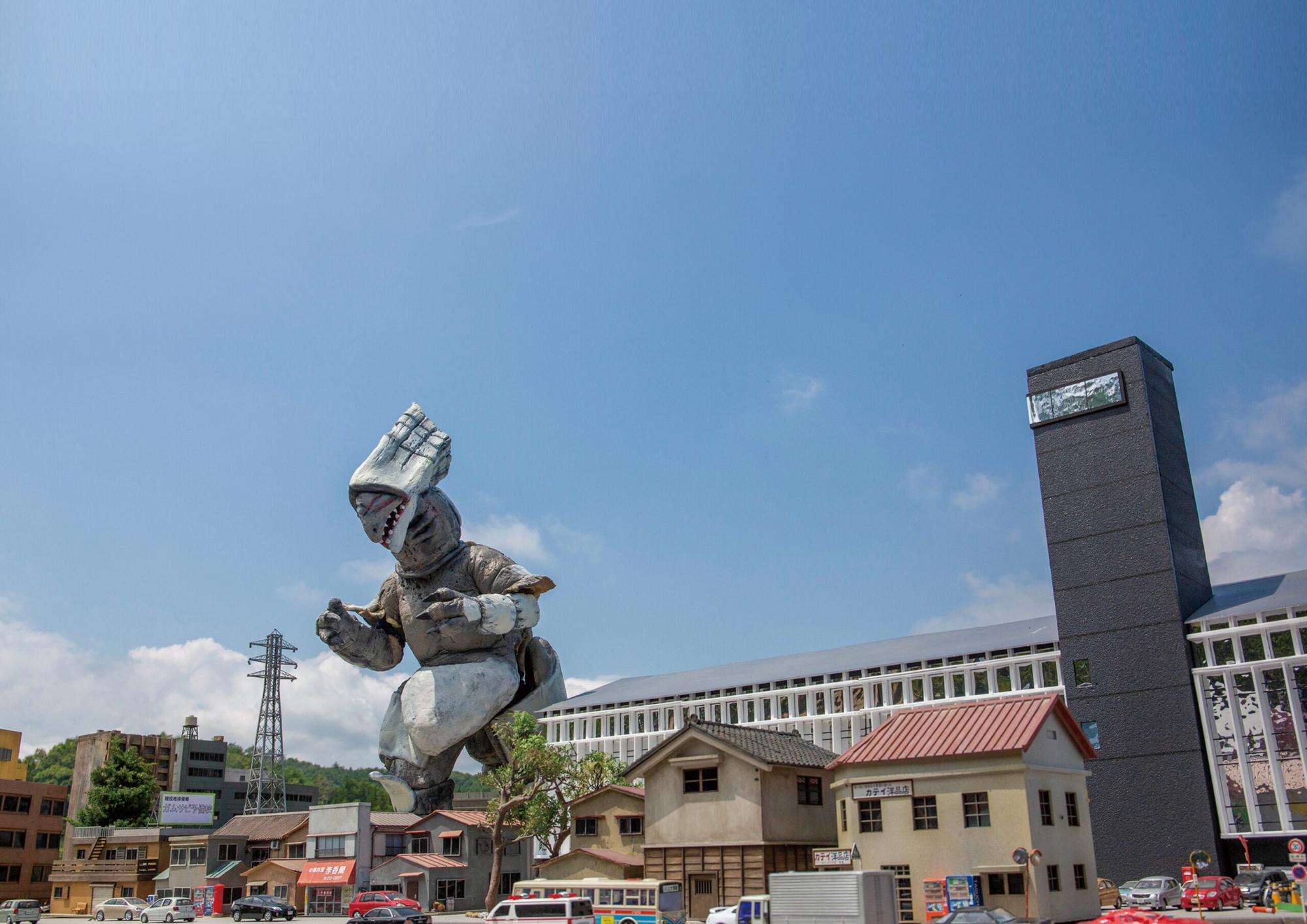 青空の下、巨大な怪獣風模型が建物の並ぶミニチュア都市に迫る迫力ある情景を捉えた写真
