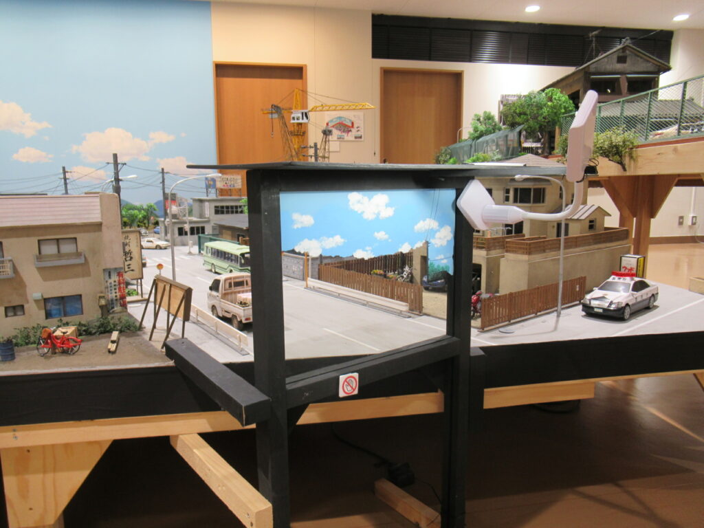 背景には青空のパネルが広がり、建物の模型や道路を走るバスや軽トラック、パトカーが配置されており、手前には黒い枠の中に雲の絵が描かれたガラスが設置されているミニチュアセットの写真