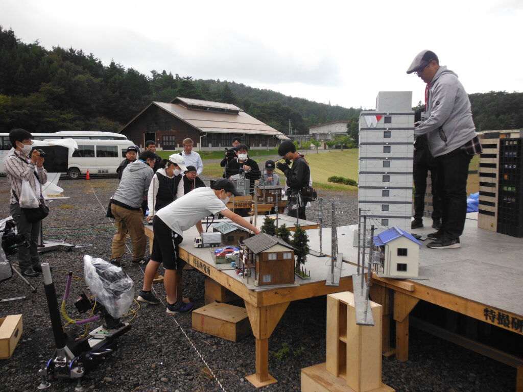 屋外に設置された特撮用のミニチュアセットの周りに大勢のスタッフや塾生が集まり、ビルや民家の模型を調整している写真