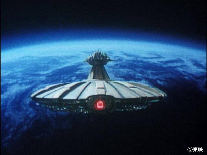 青く輝く地球の地平線を背景にして、中央に赤く光る眼のようなパーツを持つ巨大な円盤型の宇宙船が宇宙空間に浮遊しており、船体上部には細長い塔のような構造物がそびえ立っている様子を捉えた実写特撮映画の静止画