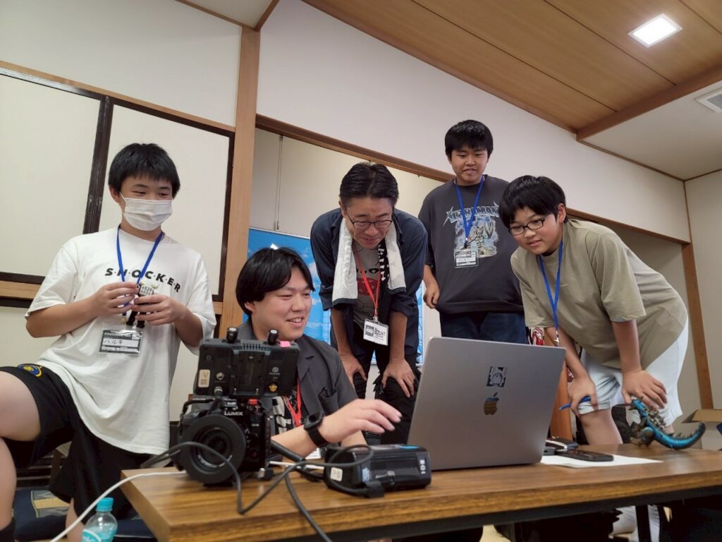 テーブル上のノートPCと撮影機材を囲んで参加者たちが画面を確認しながら議論する様子の写真