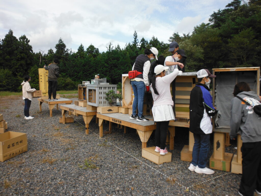 屋外の撮影現場にて、木製の土台に設置されたビルや住宅などのミニチュア模型を、スタッフや塾生が立ち上がって上部から調整したり配置を確認したりしている写真