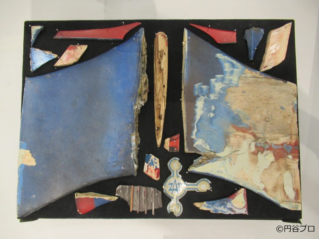 円谷プロ作品に登場する青い機体に赤い塗装が施された戦闘機の翼や機体の一部と見られる破片が、黒いボードの上に標本のように並べて固定されており、中央下部には「ZAT」のマークが描かれたエンブレム状のパーツも配置された資料写真