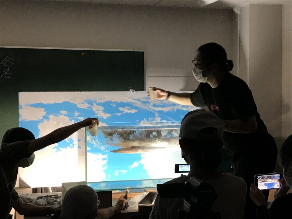 薄暗い照明の中で、青空と白い雲が描かれた背景パネルの前に置かれた透明な水槽へ、数名の参加者がコップを使って白い液体や色のついた液体を慎重に流し込み、水中で雲が広がるような独特の視覚効果を創り出している、実験的なアート制作の風景の写真