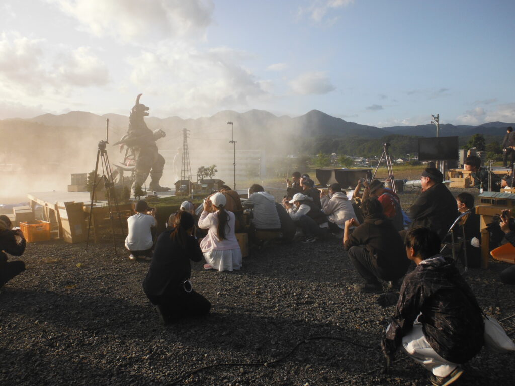 逆光で白く煙るミニチュアの街並みに怪獣が佇み、大勢のスタッフや塾生が地面にしゃがみ込みながら一斉にスマートフォンやカメラを向けて撮影している様子の写真