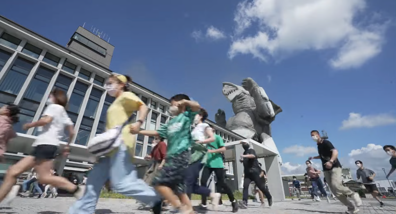 晴天の下、近代的な公共施設の建物の前で、巨大な怪獣の像を背に多くの人々が逃げ惑うように一斉に走り抜ける様子を、低いアングルから躍動感たっぷりに捉えた、特撮映画のワンシーンのような写真