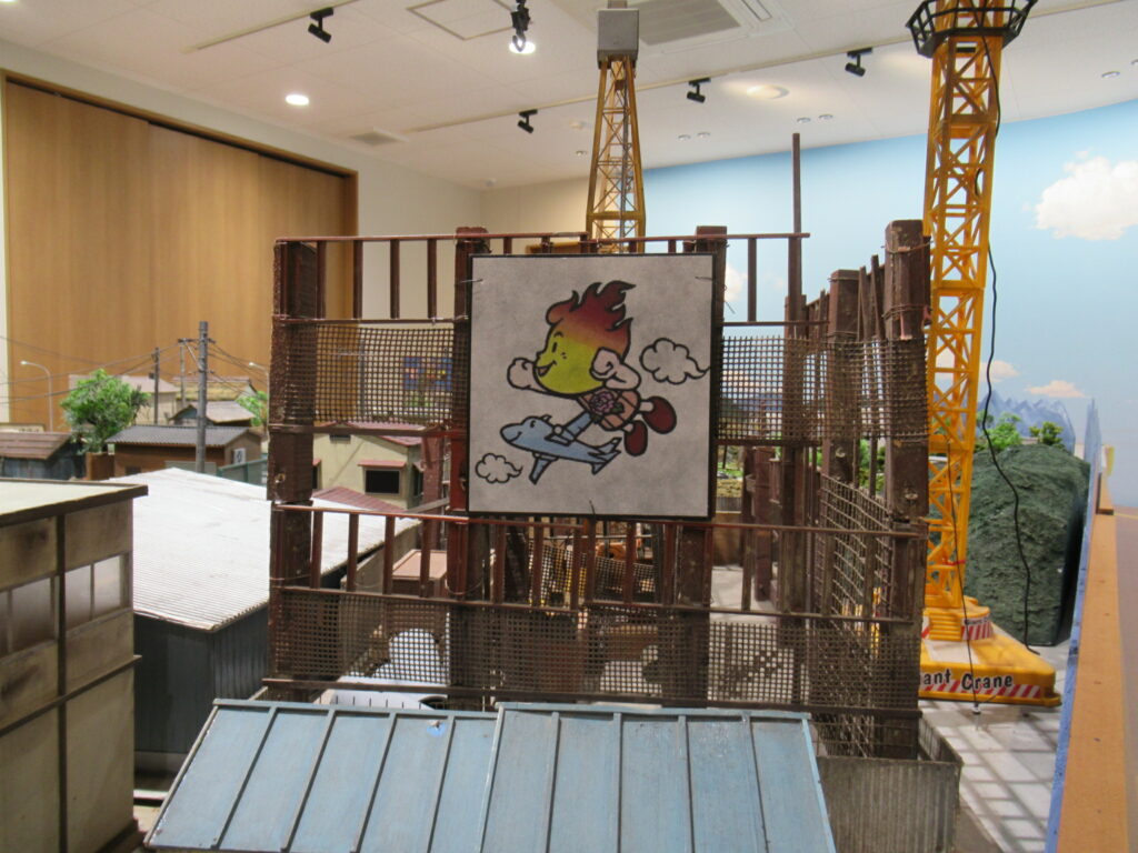 屋内の展示会場にて、ミニチュアの街並みセットを背景に、須賀川市のキャラクター「ボータン」が飛行機に乗って空を飛ぶイラストが描かれた看板が、鉄骨やネットで組まれた構造物の中央に掲げられている様子を捉えた写真