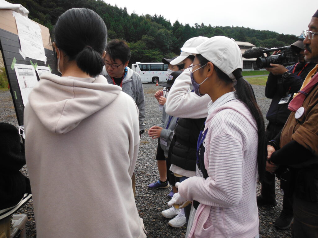 屋外の広場で、塾生やスタッフが掲示板に貼られた資料を囲んで内容を確認している写真