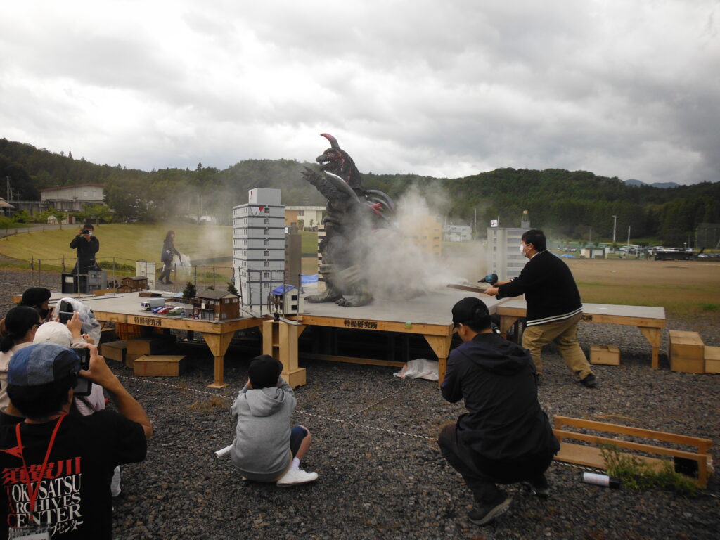 屋外に設置されたステージ上で、怪獣の模型が煙に包まれ、手前に座った塾生やスタッフたちがスマートフォンやカメラなどで撮影をしている様子の写真
