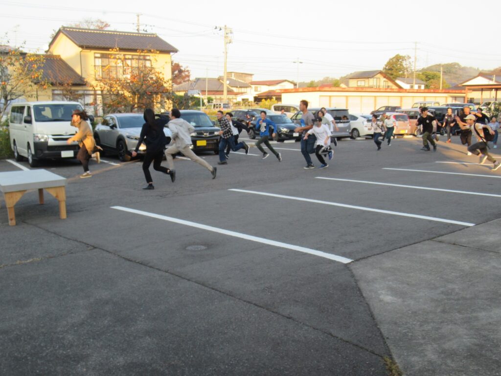 住宅地に囲まれた広い駐車場で、多くの参加者たちが駐車場を全速力で駆け抜けている、映画の避難シーンやアクションシーンの撮影を思わせる躍動感に満ちた屋外での写真
