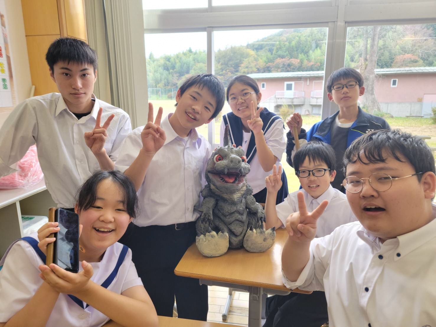 明るい教室の中で、机の上に置かれた精巧な怪獣の模型を囲み、制服姿の生徒たちが笑顔でピースサインをしたりスマートフォンを掲げたりして楽しそうにポーズをとっている、和やかな日常の風景を捉えた写真