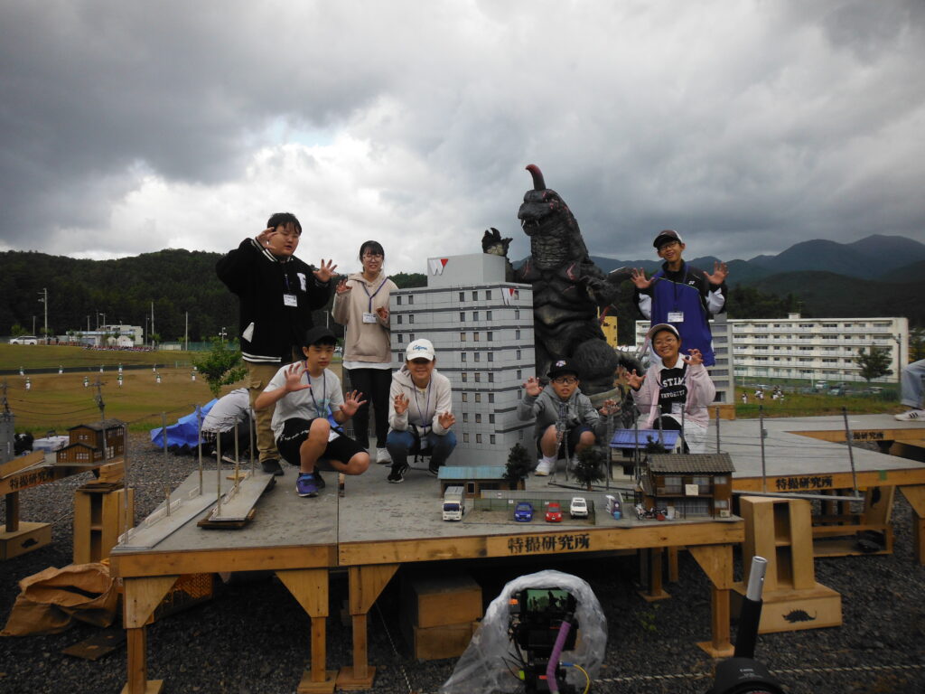 屋外に設置された特撮用のミニチュアセットの壇上に、怪獣と並んで塾生たちが集まった集合写真