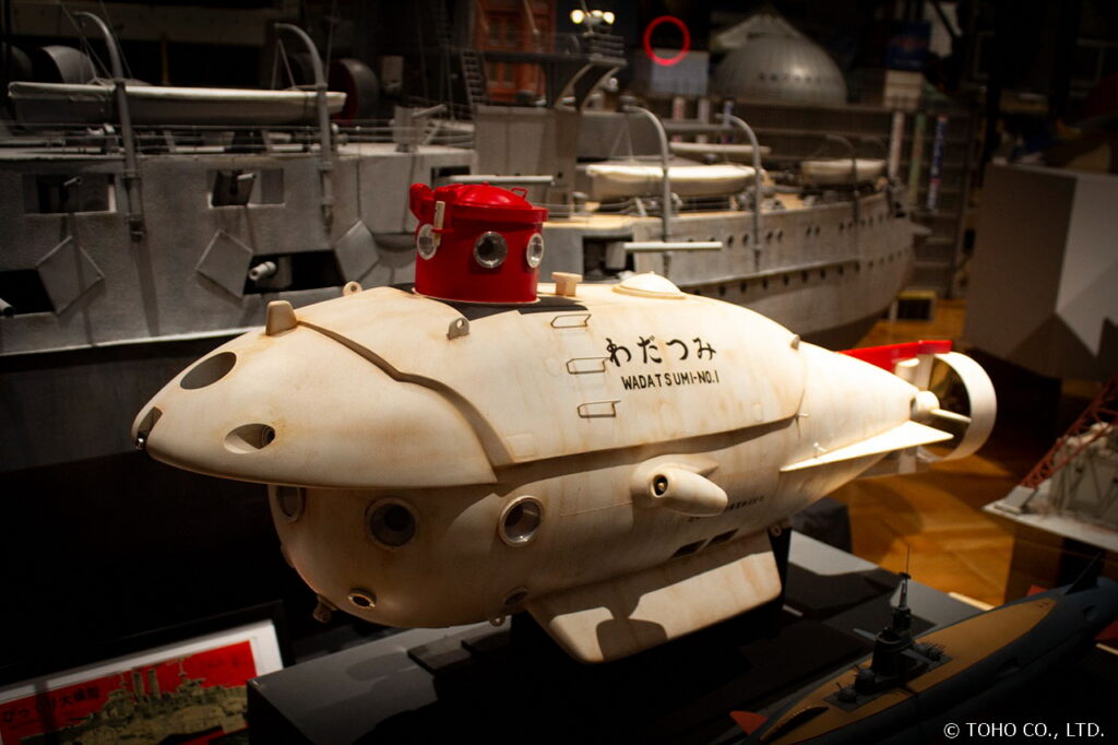 潜水艇「わだつみ」の精巧な大型ミニチュア模型が展示されており、白い機体に赤いハッチが特徴的なデザインで、背景には大きな軍艦の模型も見える、特撮映画のプロップの質感を詳細に伝える写真