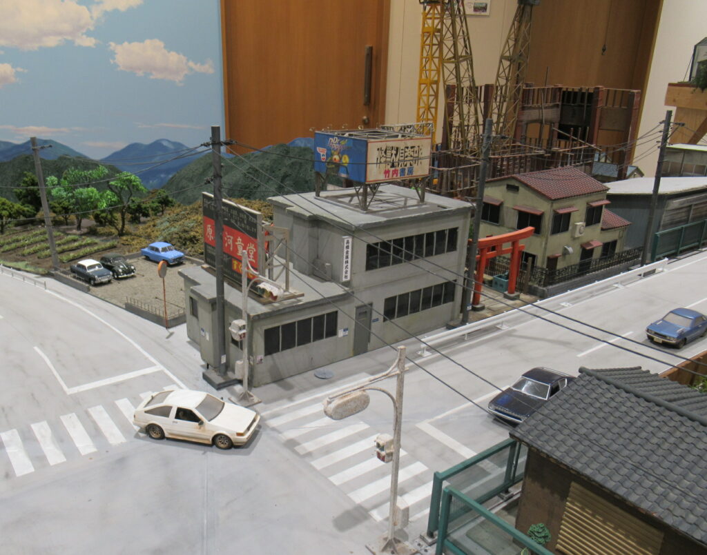 電柱や信号機が設置された交差点に白いスポーツカーなどのミニカーが並び、屋上看板のあるビルや赤い鳥居が立つ民家が路地に沿って並ぶ、昭和の街並みを精巧に再現した特撮用ミニチュアセットを斜め上から俯瞰した写真