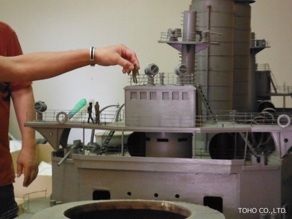 巨大な戦艦の精密なミニチュア模型の甲板部分に、人の手によって小さな人物フィギュアが置かれようとしている写真