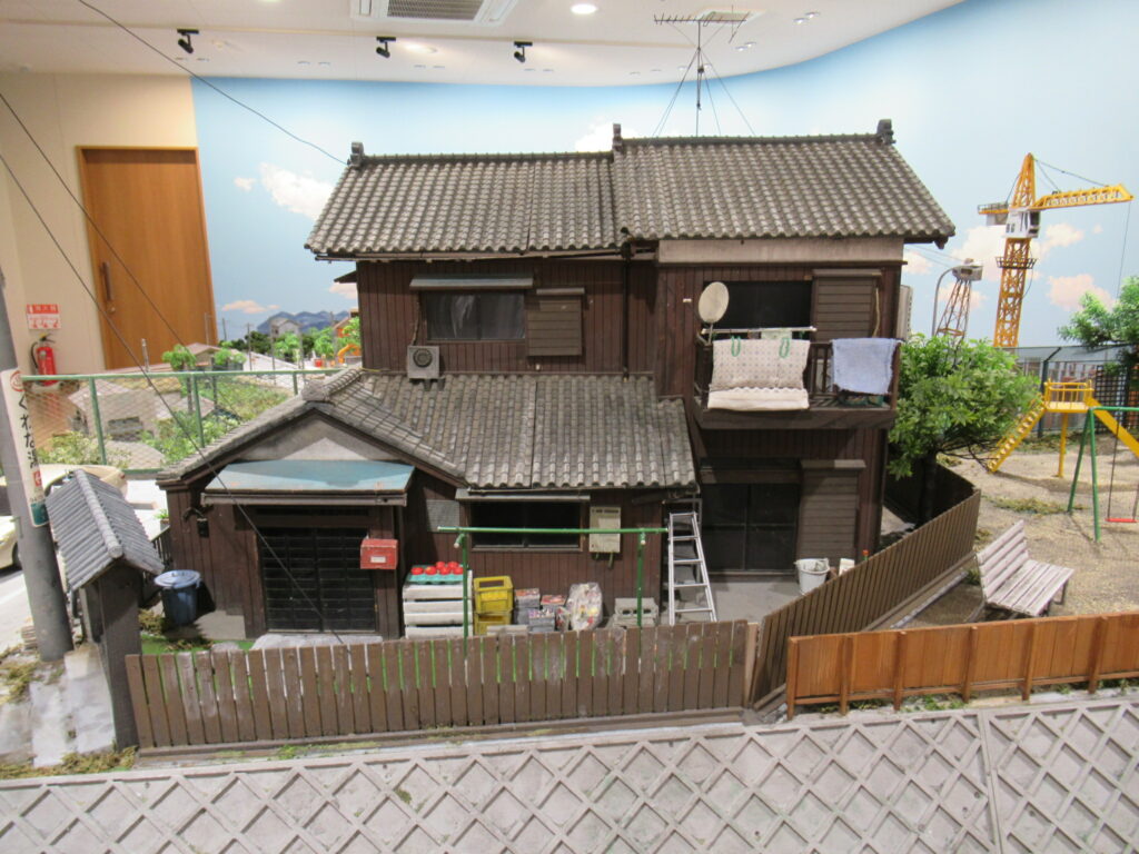 瓦屋根の二階建て民家を中心に、ベランダに干された布団や庭先の脚立、木製の柵などが細部まで作り込まれ、背景には青空の書き割りと黄色いクレーンが映り込む、生活感あふれる特撮用ミニチュアセットを正面から捉えた写真