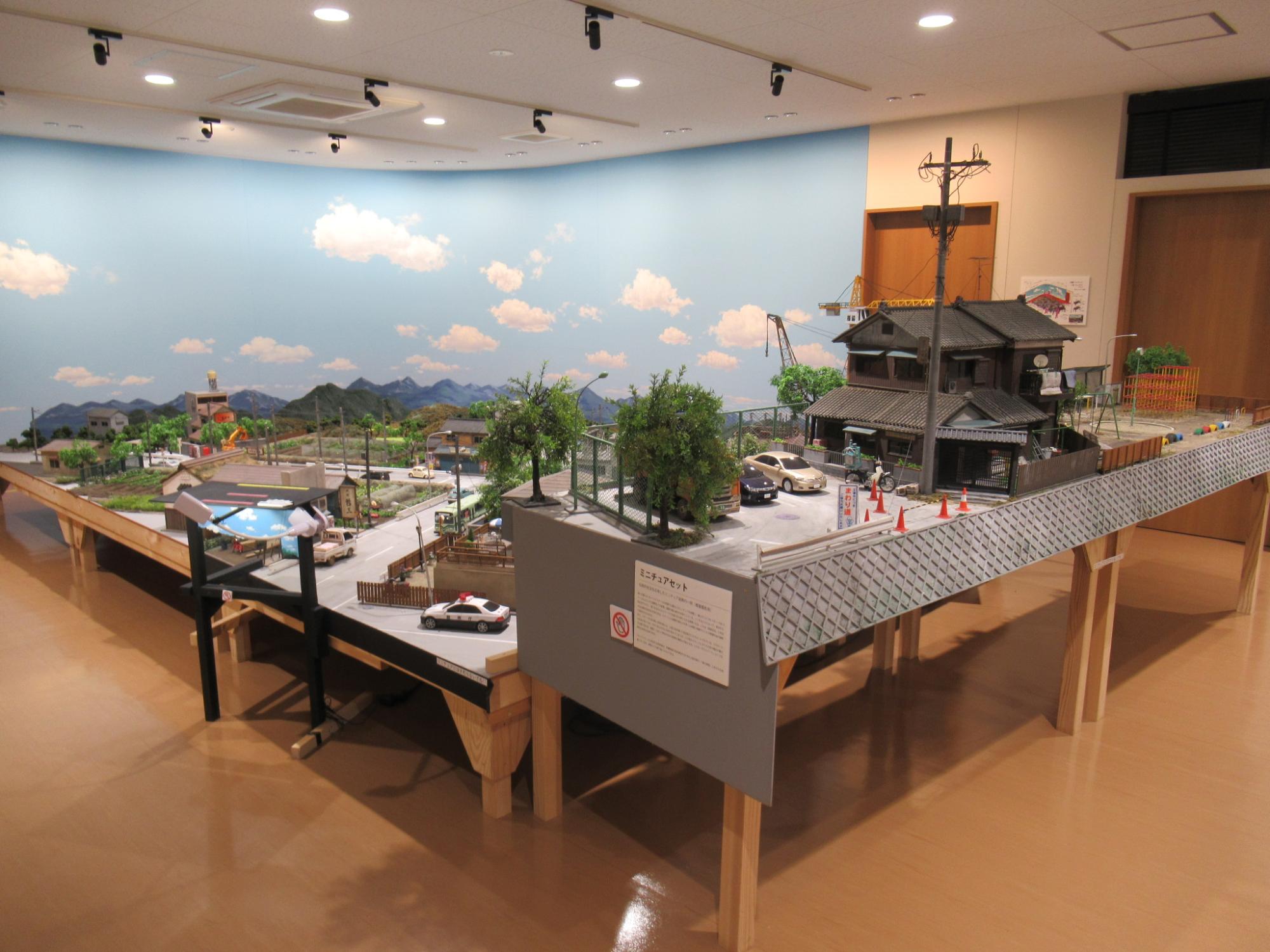 青空が描かれた背景の前に、昭和の街並みを再現した精巧なジオラマが展示されており、木造の住宅やパトカー、電柱、駐車場に並ぶミニカーなどが細部まで作り込まれた大きなミニチュアセットの写真