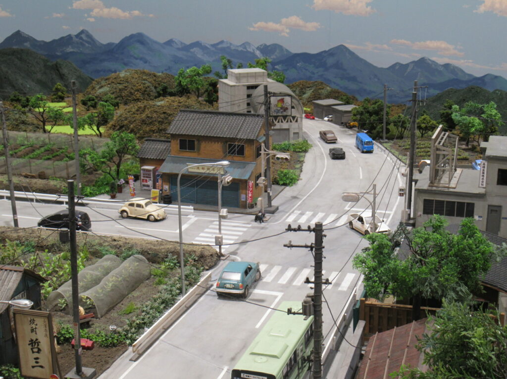 遠くに見える険しい連峰を背に、瓦屋根の民家や畑が点在する山あいの町並みを模したジオラマが広がり、道路上の交差点にはバスや乗用車が精密に配置され、のどかな地方の風景が細部まで再現されているミニチュア写真