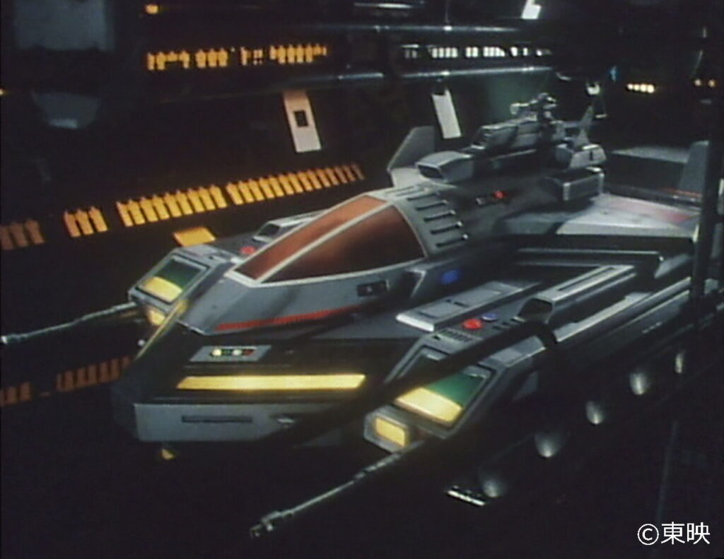 暗い格納庫のような場所で、機体各部のライトやパネルを黄色く発光させながら、今まさに出撃の時を待つように静かに鎮座している、黒とグレーを基調とした重厚で近未来的なデザインの特殊戦闘車両を捉えた当時の特撮作品の写真