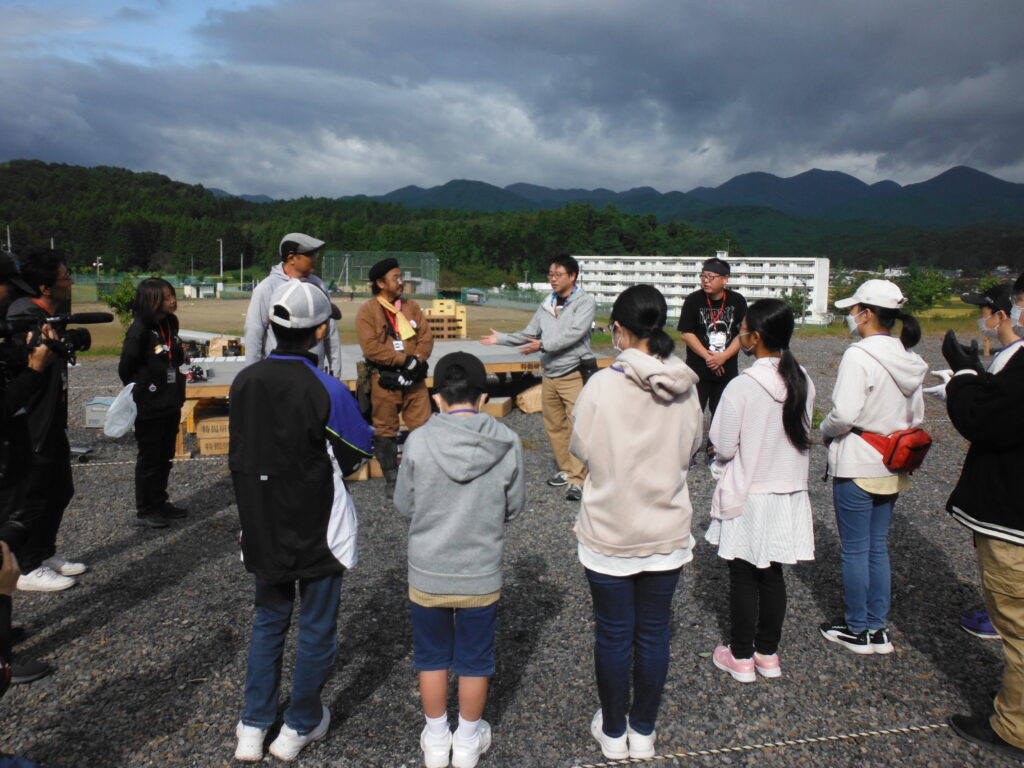 屋外の砂利敷きの広場で、講師が身振り手振りを交えて話し、それを囲むようにして話を聞く塾生たちの様子を背後から写した写真