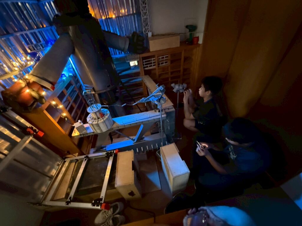 暗い室内で発光する巨大ロボット風セットの前で二人が装置や模型を操作し撮影準備を進める写真