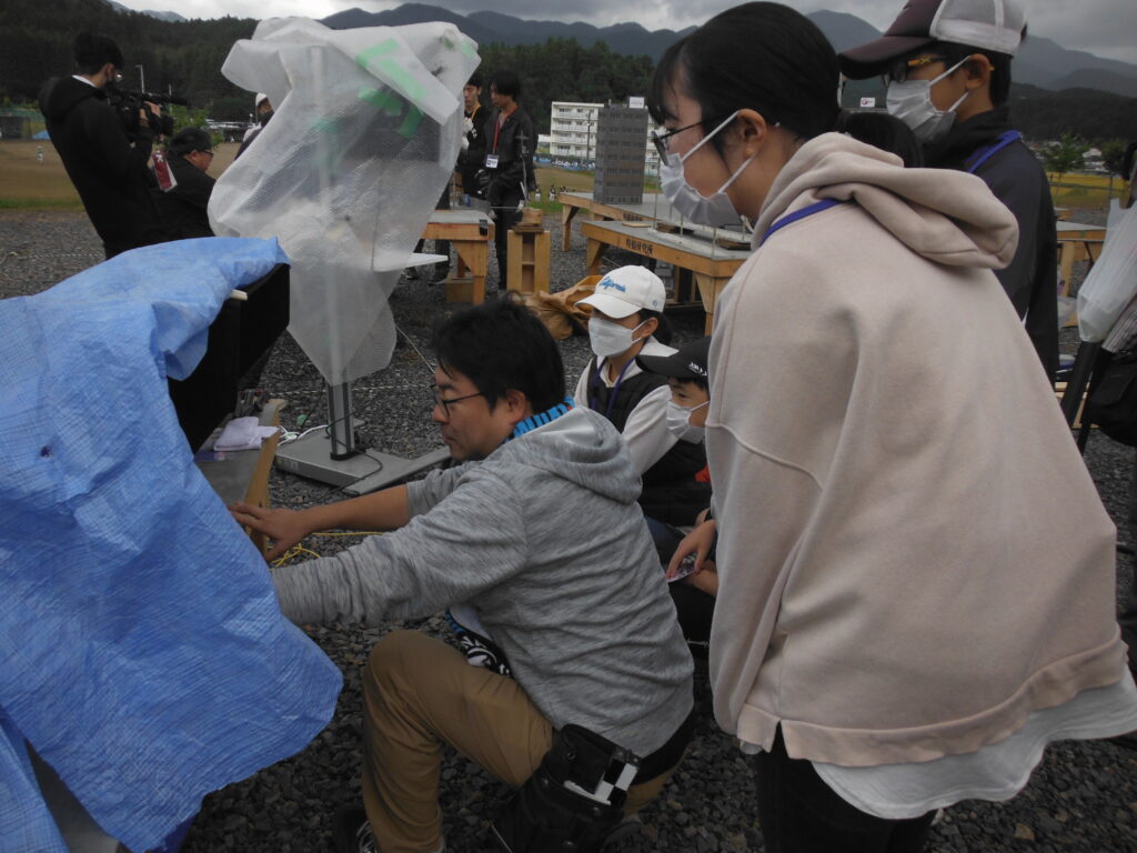 砂利敷きの広場で、ブルーシートや白い保護カバーがかけられた撮影機材の周りに、塾生が集まり説明を間近で聞いている様子の写真