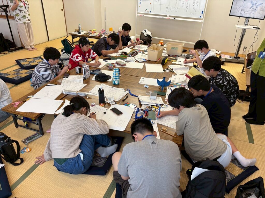 畳敷きの部屋で大きな座卓を囲む参加者たちが紙や資料や道具を広げて熱心に創作作業へ取り組む写真