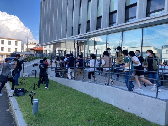 須賀川市役所の外にあるスロープで、カメラを構えたスタッフが見守る中、多くの人々が何かに驚いたり逃げたりするかのように一斉に駆け抜けていく、躍動感のある撮影風景を捉えた写真