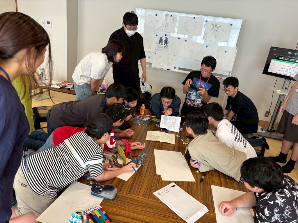 テーブルに集まった子どもたちが怪獣フィギュアや資料を囲んで創作作業に取り組む様子の写真