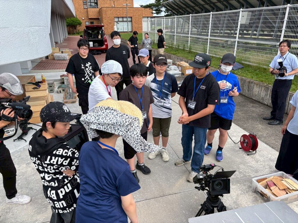 屋外で撮影機材や木材が並ぶ中、参加者とスタッフがカメラ前に集まり打ち合わせを行う写真