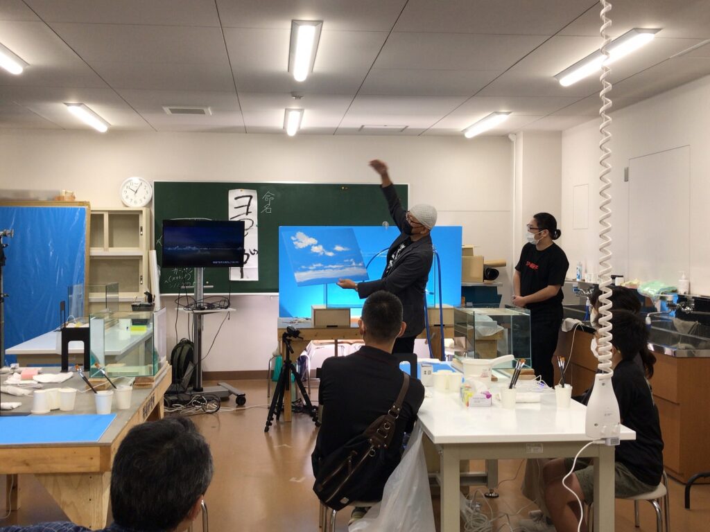 明るい教室の中で、眼鏡をかけた講師の男性が青空と白い雲が描かれた絵画を掲げながら身振り手振りを交えて熱心に解説しており、それを見守る数名の参加者や机に並んだ画材から、絵画教室での活発な学びの様子が伝わる写真
